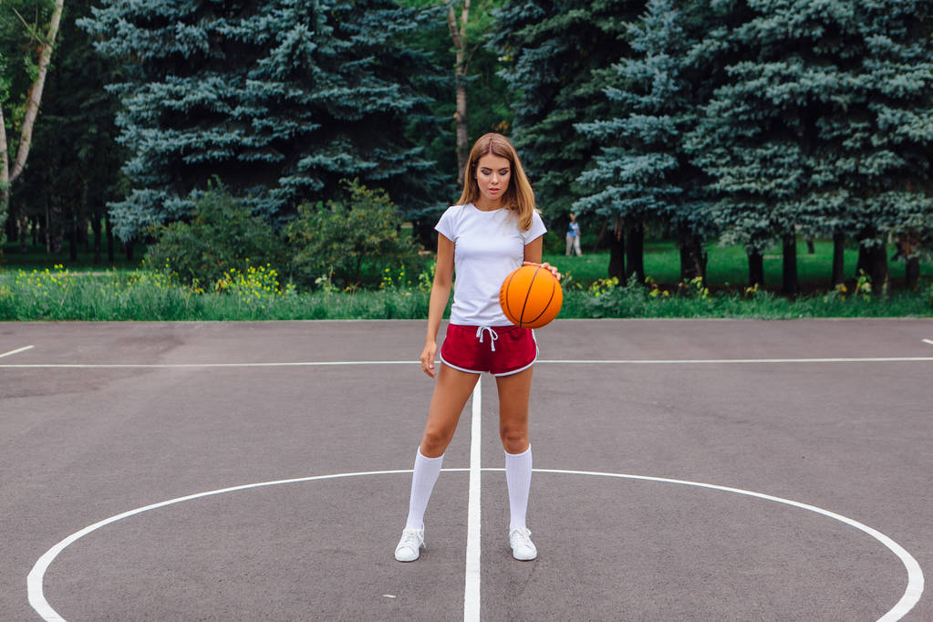 白の t シャツ、ショート パンツ、スニーカー、服を着た美しい少女はバスケット ボール コートでボールで遊ぶ. - 写真・画像
