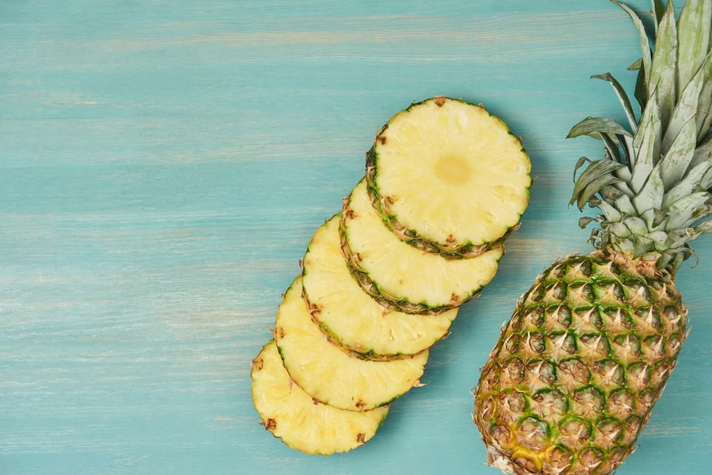 Draufsicht auf geschnittene saftige Kreise in der Nähe ganzer Ananas auf türkisfarbenem Holztisch - Foto, Bild