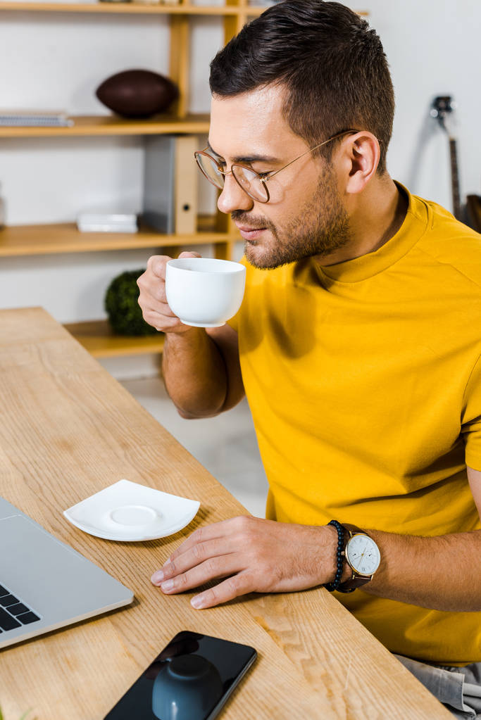 hombre guapo oliendo café mientras está sentado en el escritorio
 - Foto, Imagen