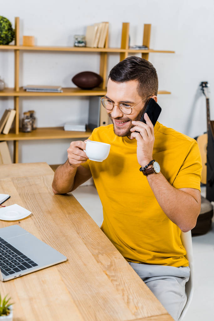 hombre guapo en gafas hablando en el teléfono inteligente y sosteniendo la taza de café
 - Foto, imagen