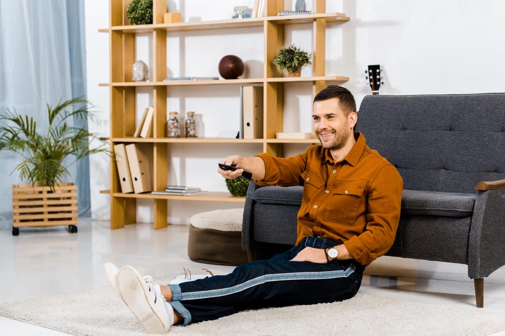 hombre alegre sosteniendo el control remoto y sentado en el suelo en la sala de estar moderna
  - Foto, Imagen