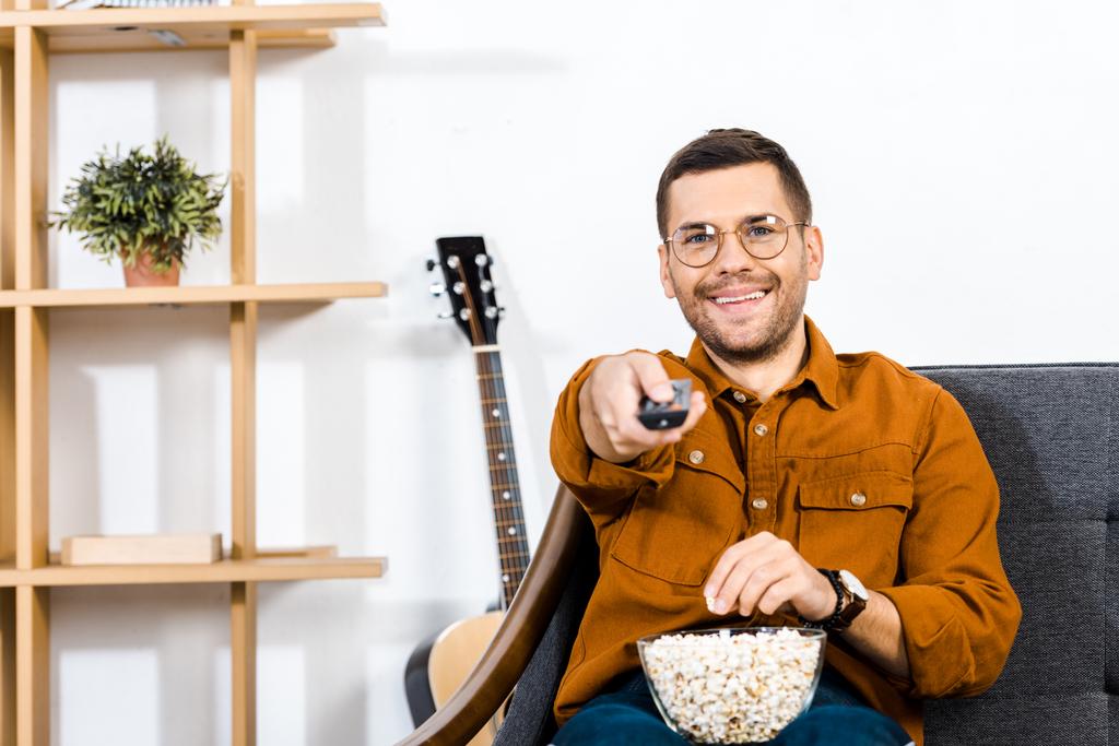iloinen mies istuu sohvalla popcorn kulhoon ja tilalla kaukosäädin
  - Valokuva, kuva