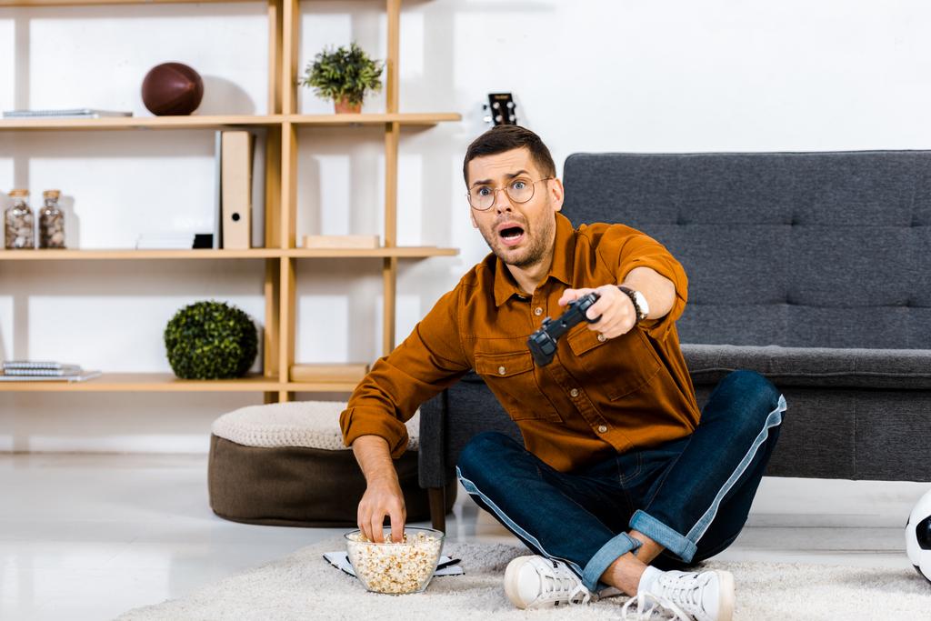 zszokowany mężczyzna oglądanie telewizji i siedząc w pobliżu bowl popcorn  - Zdjęcie, obraz