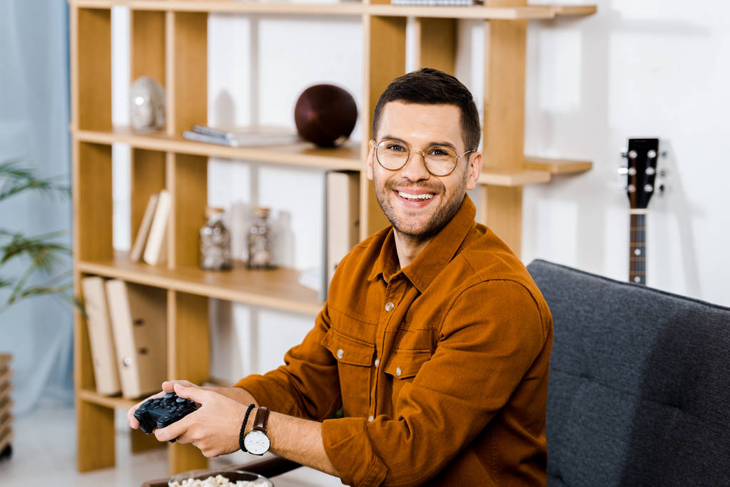 vrolijke man in glazen houden gamepad in woonkamer - Foto, afbeelding