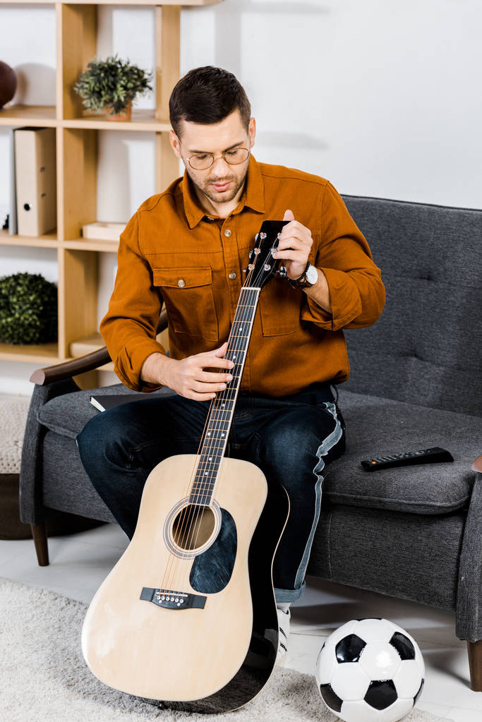 όμορφος άντρας με τα γυαλιά, κάθεται στον καναπέ και tuning ακουστική κιθάρα στο σπίτι - Φωτογραφία, εικόνα