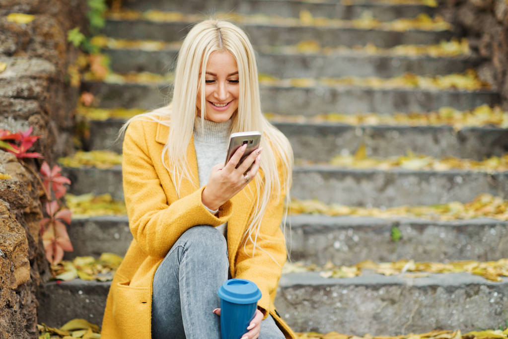 Mujer rubia joven sentada en el parque de la ciudad y tomando café para llevar, usando teléfono móvil
 - Foto, Imagen