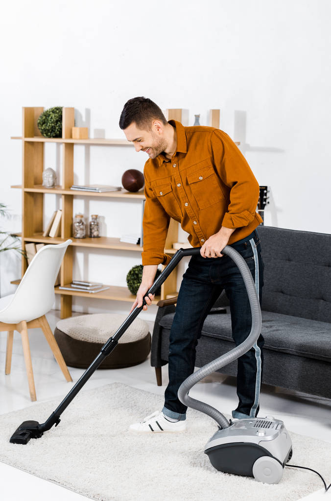 knappe man in moderne woonkamer glimlachend en het reinigen van huis met hoover  - Foto, afbeelding