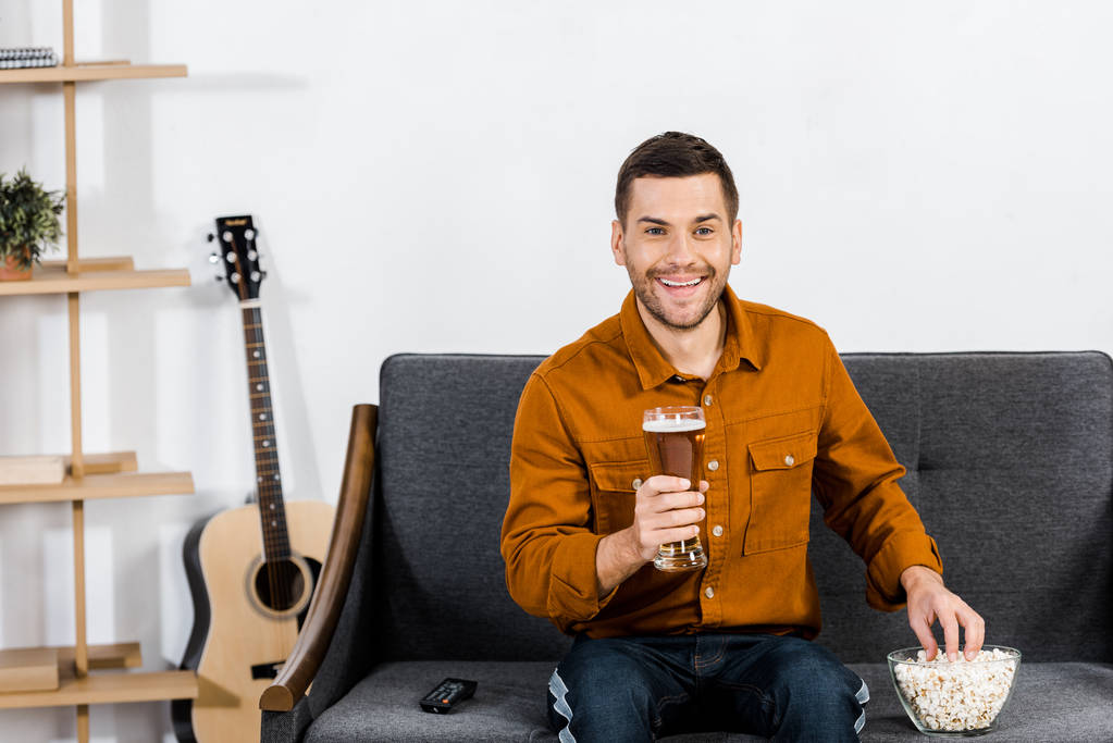 hombre guapo en la sala de estar moderna sosteniendo un vaso de cerveza y comiendo palomitas de maíz
 - Foto, Imagen