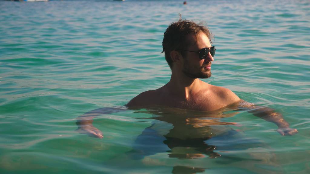 Portret van een knappe jonge man met een baard in zonnebril ontspannen in kristalhelder zeewater - Foto, afbeelding