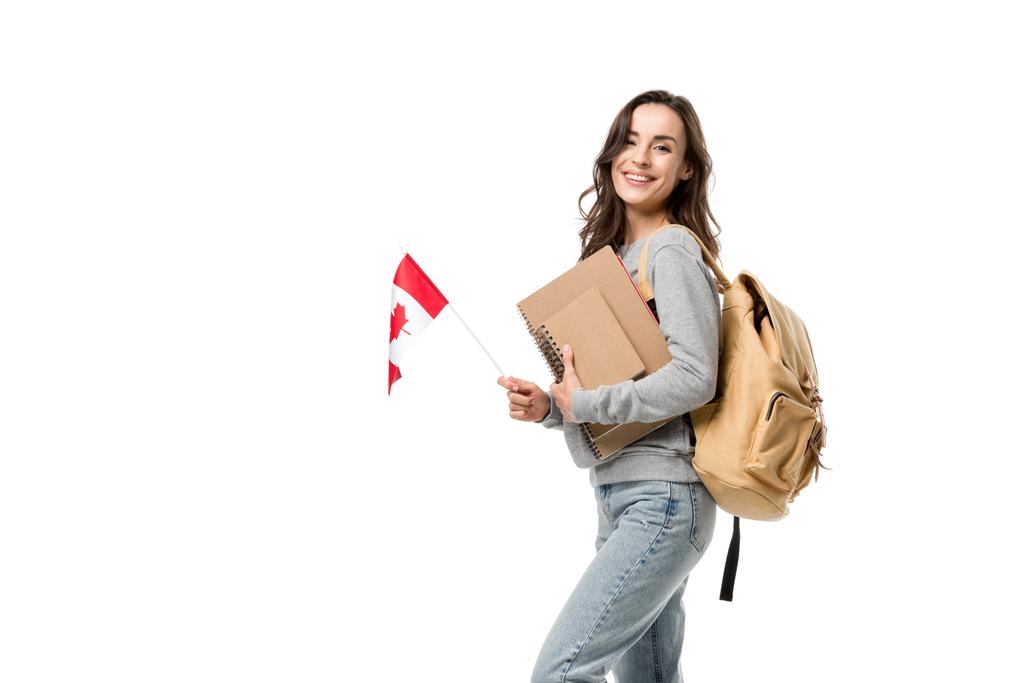 glückliche Studentin mit Rucksack und Notizbüchern, die in die Kamera blickt, während sie die kanadische Flagge isoliert auf weiß hält - Foto, Bild