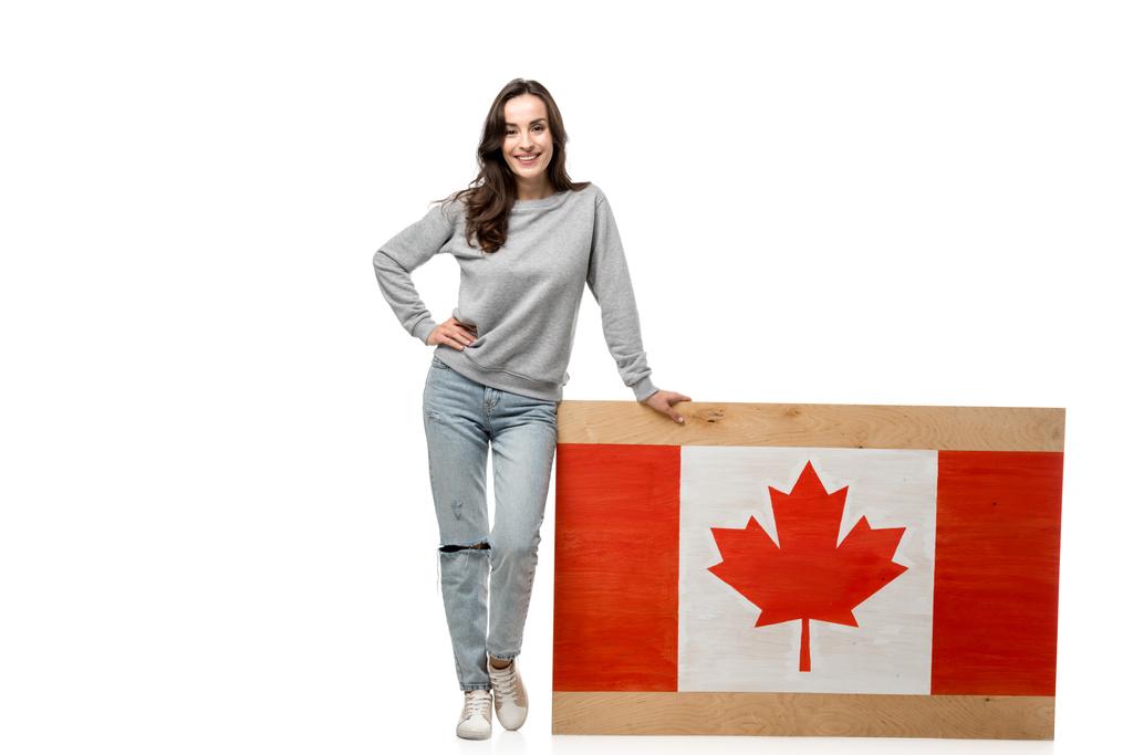χαμογελαστό γυναίκα με το χέρι στο ισχίο στέκεται κοντά σε ξύλινη σανίδα με καναδική σημαία που απομονώνονται σε λευκό - Φωτογραφία, εικόνα