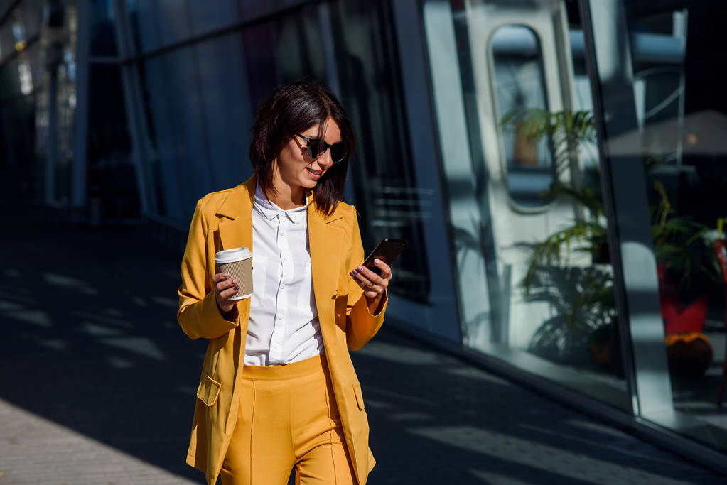 Kaukázusi, fiatal üzleti nő használ szúró telefon és italok kávé kívül az office-központ közelében. Vonzó nő görgetés oldalak, megérinti a touchscreen, Hírek és pénzügyi térképek ellenőrzése. - Fotó, kép