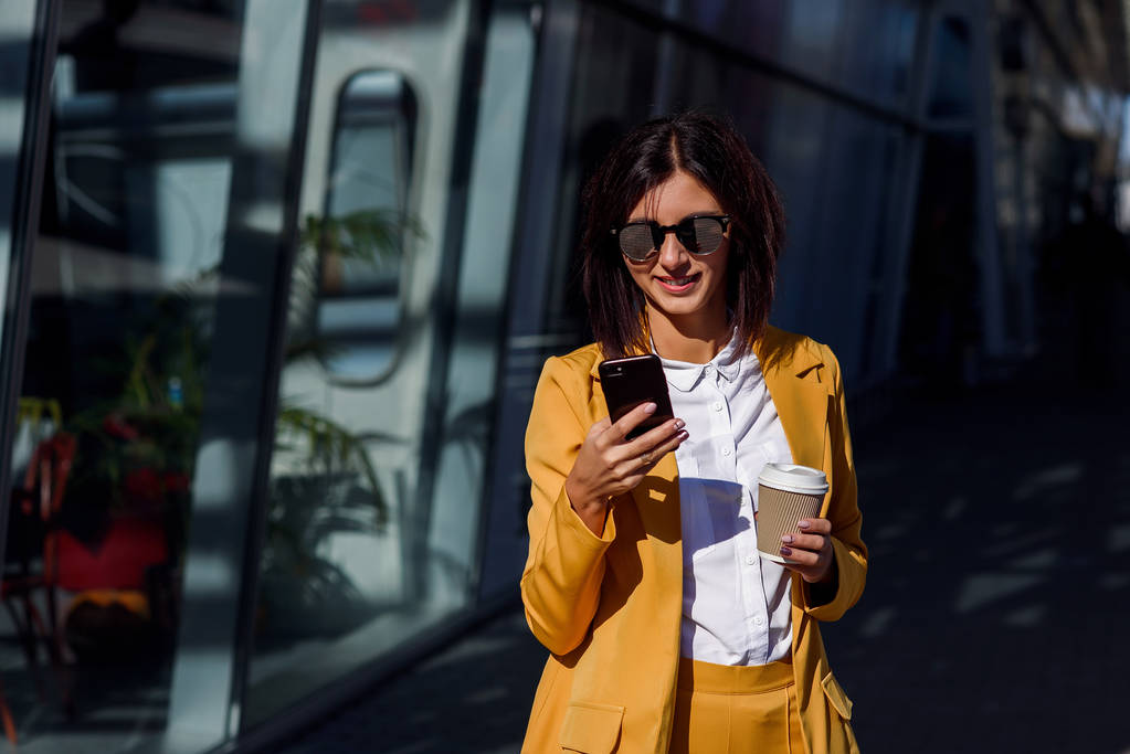 Кавказская молодая деловая женщина пользуется смартфоном и пьет кофе на улице возле офисного центра. Привлекательная женщина прокручивает страницы, нажимает на сенсорный экран, проверяет новости и финансовые графики
. - Фото, изображение