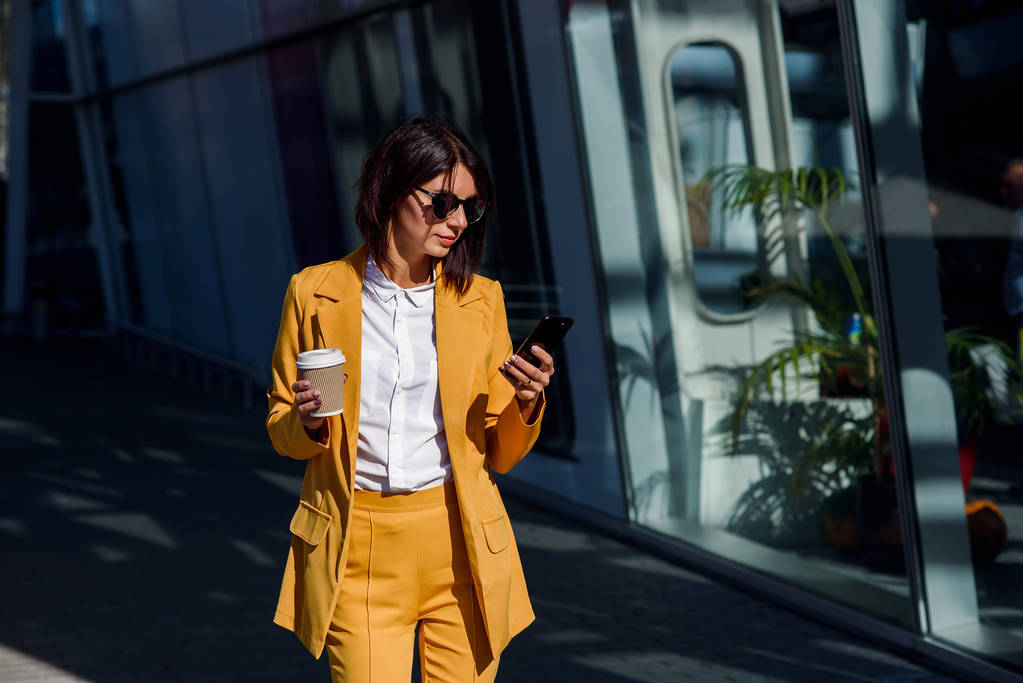 Eine kaukasische junge Geschäftsfrau nutzt ihr Smartphone und trinkt draußen in der Nähe des Bürozentrums Kaffee. attraktive Frau blättert Seiten, tippt auf Touchscreen, checkt Nachrichten und Finanzcharts. - Foto, Bild