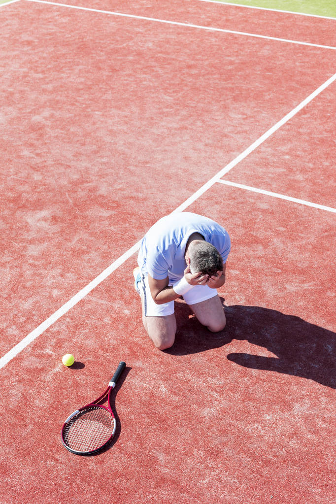 Comprimento total de homem maduro desapontado com a cabeça nas mãos enquanto se ajoelha por raquete de tênis na quadra vermelha durante o verão
 - Foto, Imagem