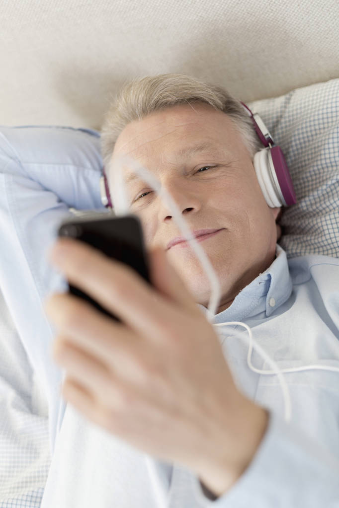 Kypsä mies katselee älypuhelinta makatessaan sängyssä kotona
 - Valokuva, kuva