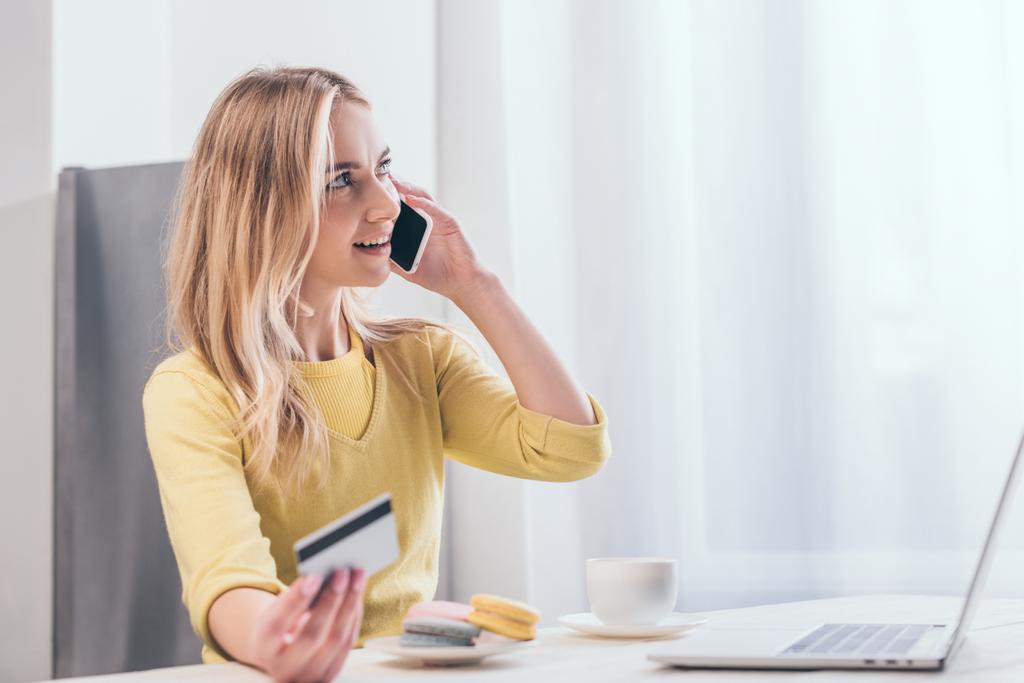 atractiva mujer rubia hablando en el teléfono inteligente mientras sostiene la tarjeta de crédito cerca de la computadora portátil
 - Foto, imagen