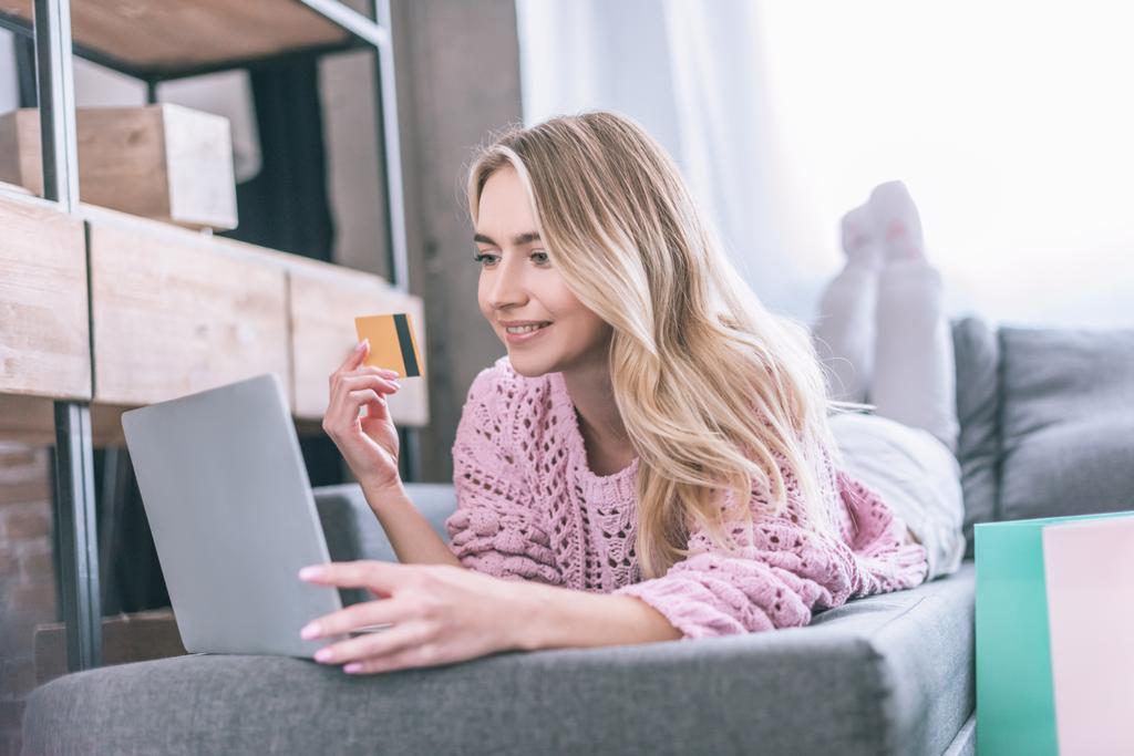 smiling woman holding credit card while lying on sofa with laptop - Photo, Image