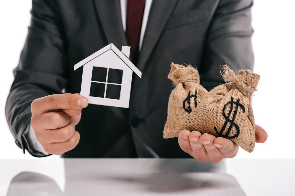 обрезанный вид ипотечного брокера, держащего бумажный дом и мешки с деньгами изолированные на белом
 - Фото, изображение