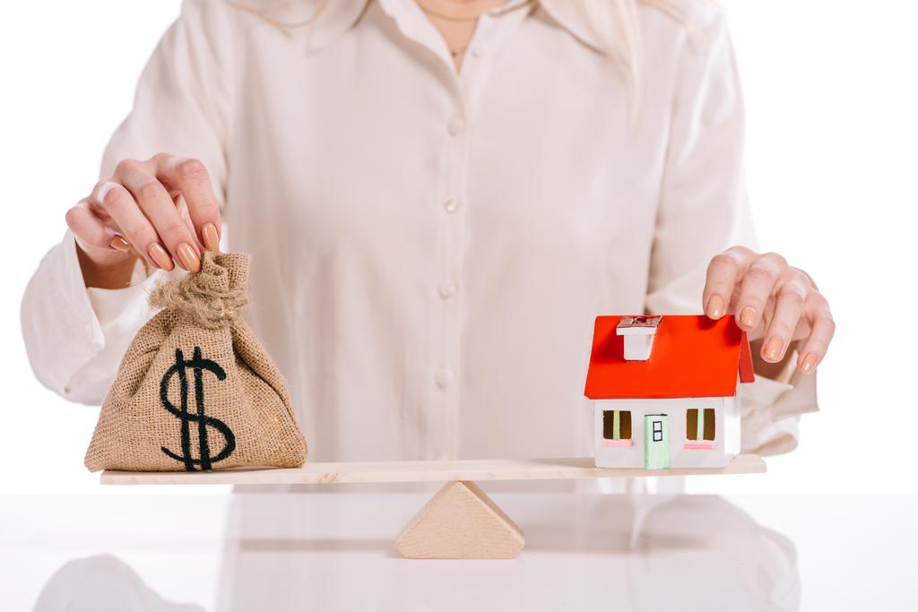 обрезанный взгляд бизнесвумен взвешивая модель дома и мешок денег изолированы на белом, ипотечная концепция
 - Фото, изображение