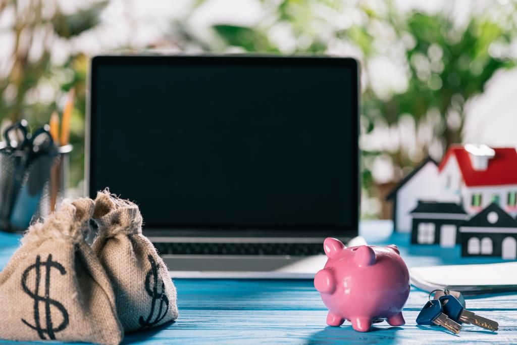 Selektywny fokus moneybags, pink piggy bank i klucze na drewnianym stole z laptopa na tle, koncepcja kredyt hipoteczny - Zdjęcie, obraz