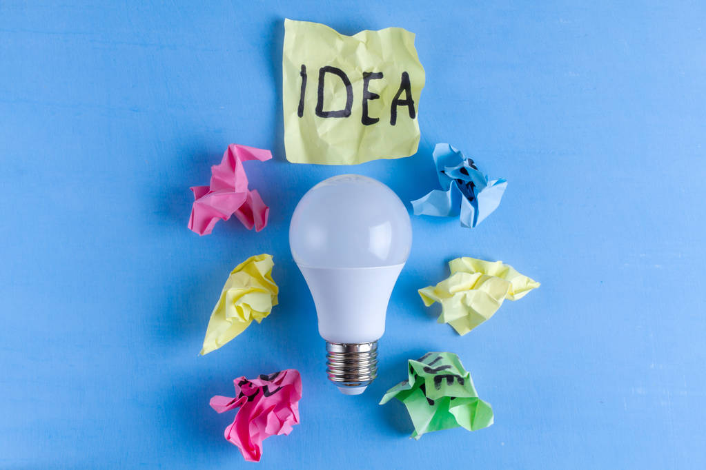 Ideia conceito. Uma ideia nova. Bolas de papel amassadas coloridas, caderno e lâmpada sobre um fundo azul. Comecem. Espaço de cópia
  - Foto, Imagem