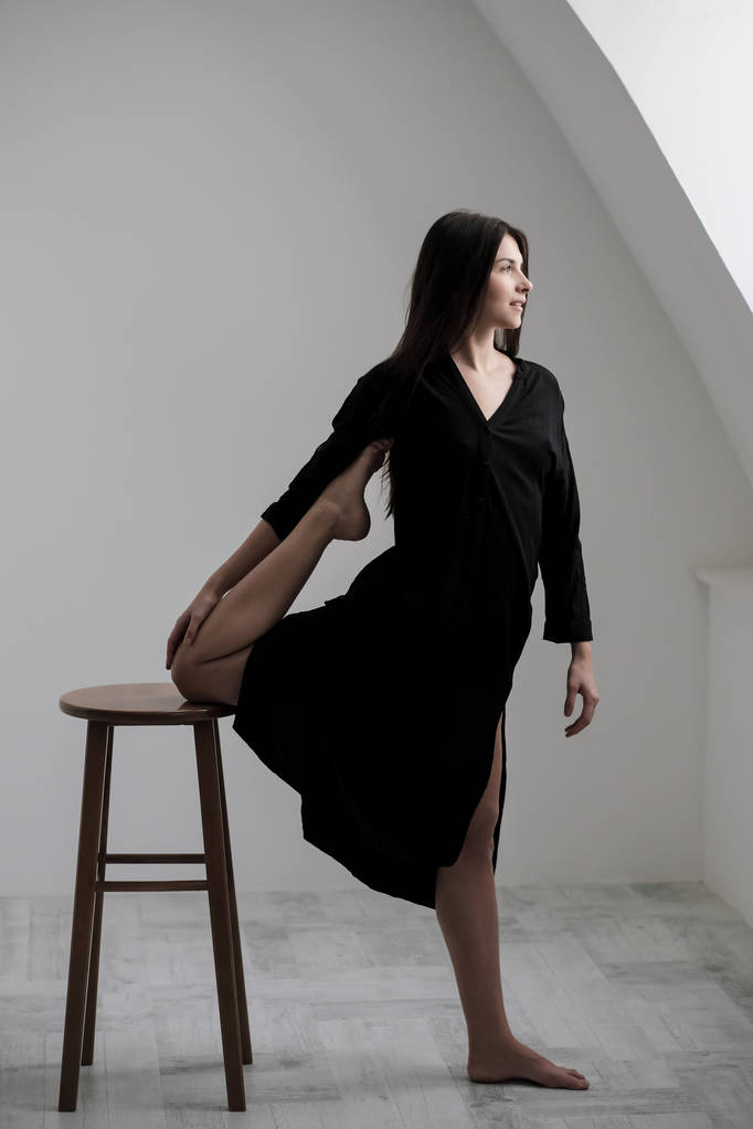 Eine junge Tänzerin im schwarzen Kleid streckt sich am Fenster. Weiße Halle, Arbeiterklasse, Yoga-Asanas. - Foto, Bild