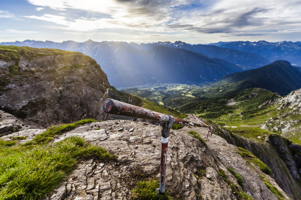 Mahtava alppimaisema, jossa on käsijohde vaelluspolulla ja auringonsäteitä laaksossa
 - Valokuva, kuva