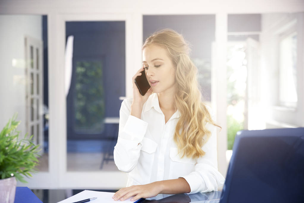 Портрет красивой молодой деловой женщины, сидящей за рабочим столом и разговаривающей с кем-то по мобильному телефону
 - Фото, изображение