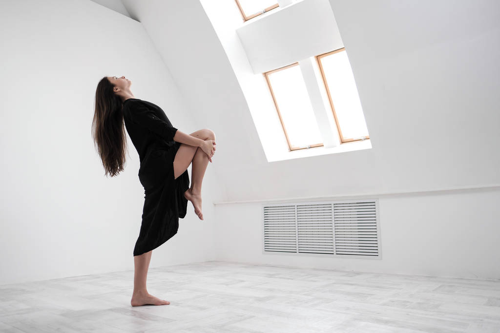 Een jonge danseres in een zwarte jurk is uitrekken zich bij het raam. White Hall, de werkende klasse, yoga asana 's. - Foto, afbeelding