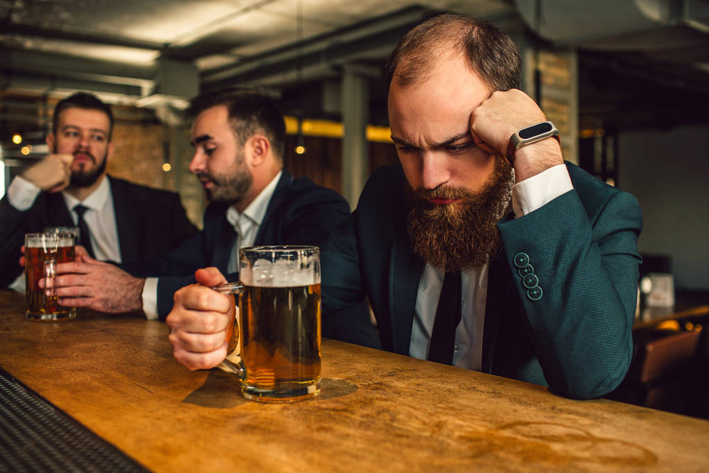 Young man in suit sit and sleep. He prop up head. Guy hold beer mug. Other two office workers sit behind and talk. - Photo, Image