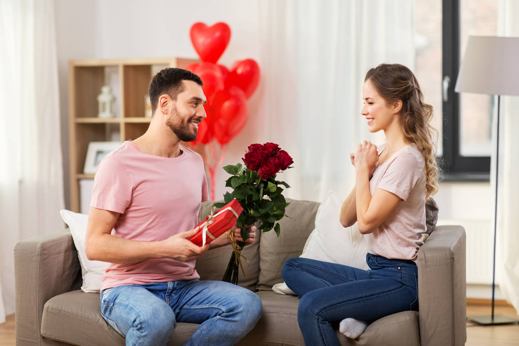 día de San Valentín, pareja, relaciones y concepto de personas - hombre feliz dando flores a la mujer y presente en casa
 - Foto, imagen