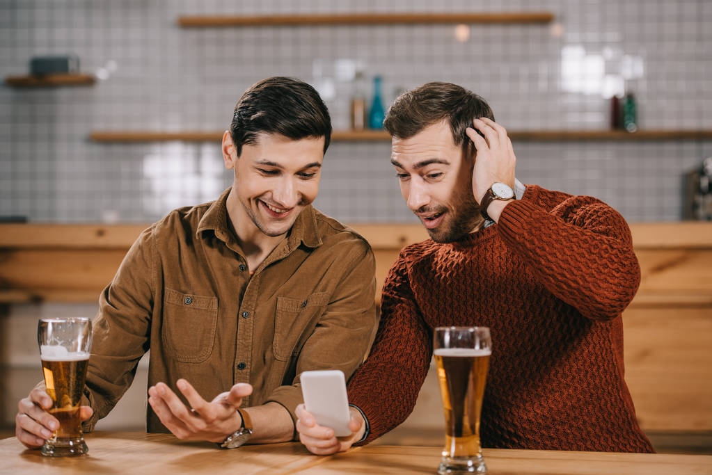 удивленные мужчины смотрят на смартфон рядом со стаканами с пивом
 - Фото, изображение
