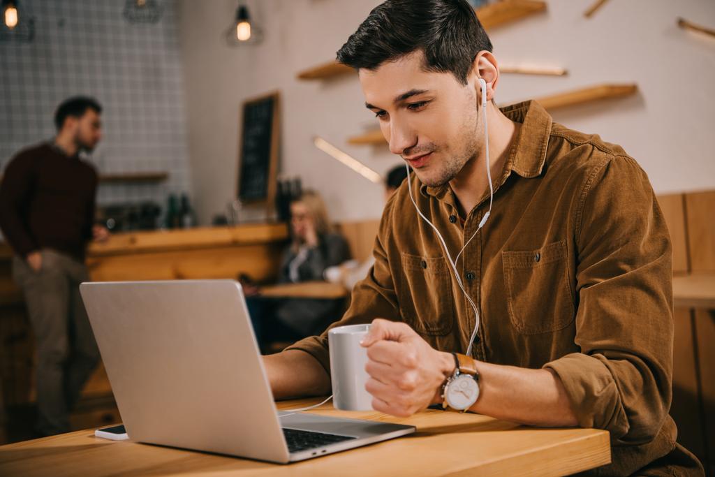 homem bonito em fones de ouvido segurando xícara de café e olhando para laptop no café
 - Foto, Imagem