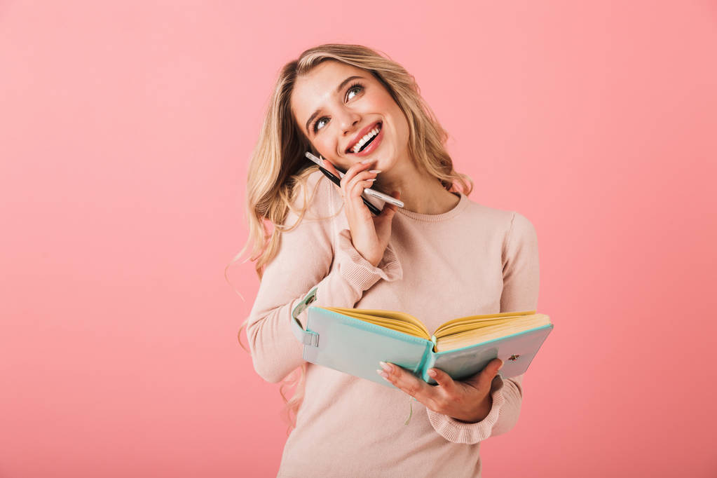 Retrato de una joven alegre con suéter de pie aislado sobre fondo rosa, escribiendo notas en un diario mientras habla por teléfono móvil
 - Foto, Imagen