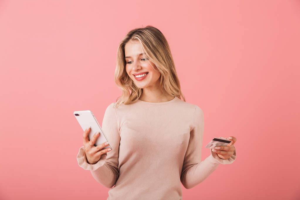 Portrait of a happy young woman wearing sweater standing isolated over pink background, using mobile phone, showing credit card - Photo, Image