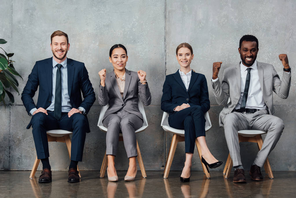 улыбающиеся многонациональные бизнесмены, сидящие на стульях и аплодирующие сжатыми кулаками в зале ожидания
 - Фото, изображение