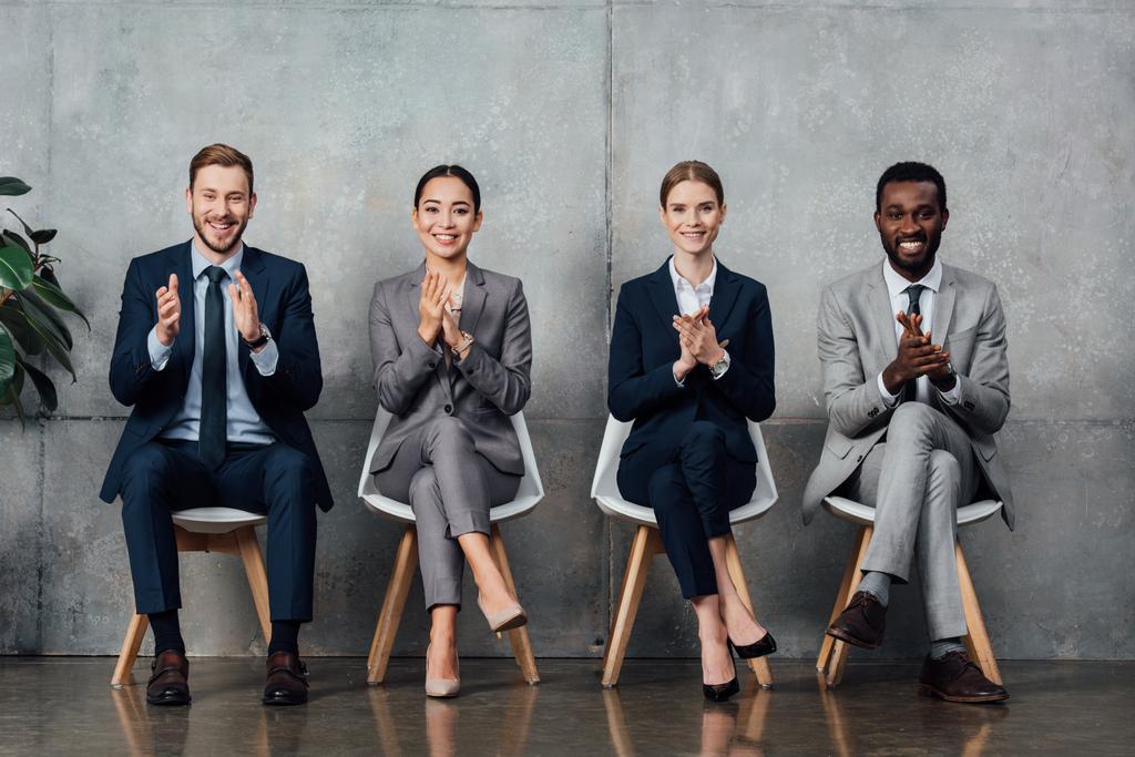 felices empresarios multiétnicos sentados en sillas y aplaudiendo en la sala de espera
 - Foto, Imagen