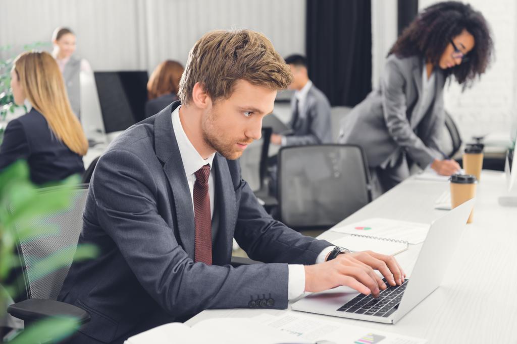 joven empresario concentrado que utiliza el ordenador portátil mientras trabaja con colegas en la oficina de espacio abierto
  - Foto, imagen
