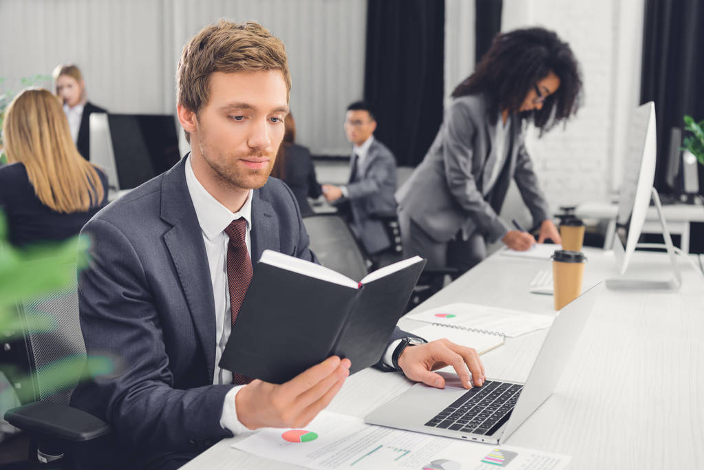 jeune homme d'affaires concentré tenant un bloc-notes et utilisant un ordinateur portable dans un bureau ouvert
 - Photo, image