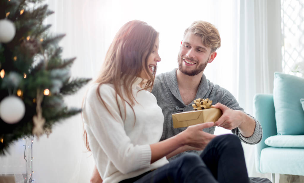 Happy couple are giving themselves Christmas gifts sitting on floor next to Xmas tree - Photo, Image