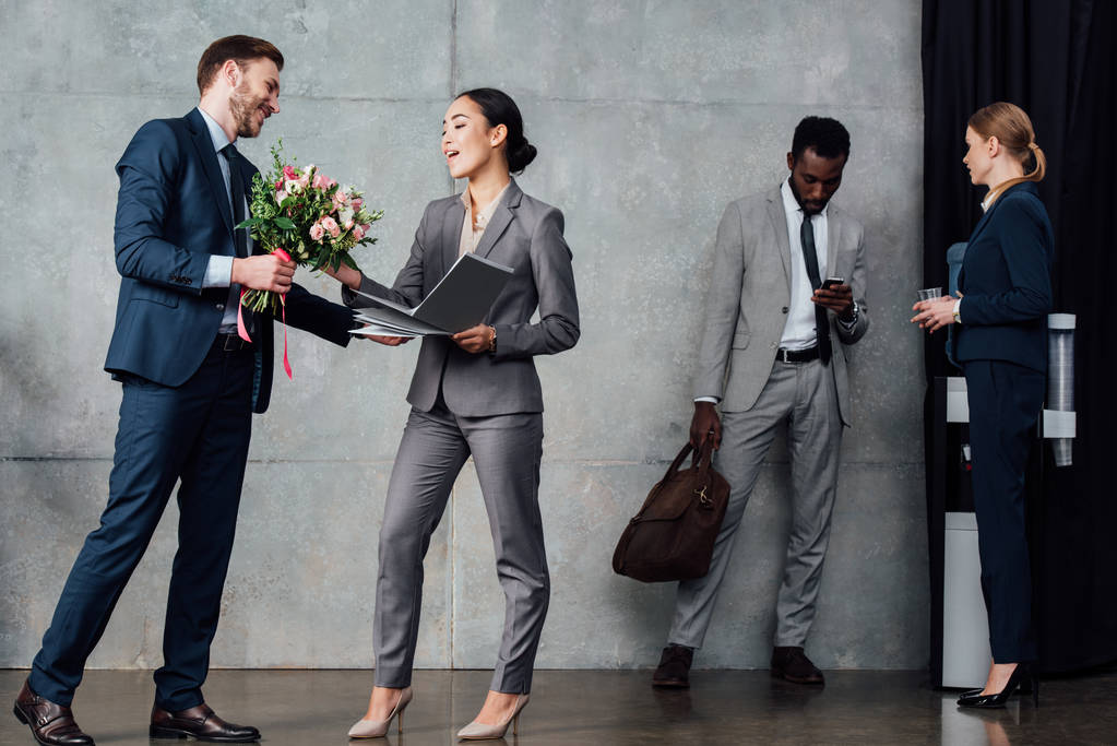 uomo d'affari che presenta fiori a donna d'affari con colleghi multietnici sullo sfondo in sala d'attesa
 - Foto, immagini
