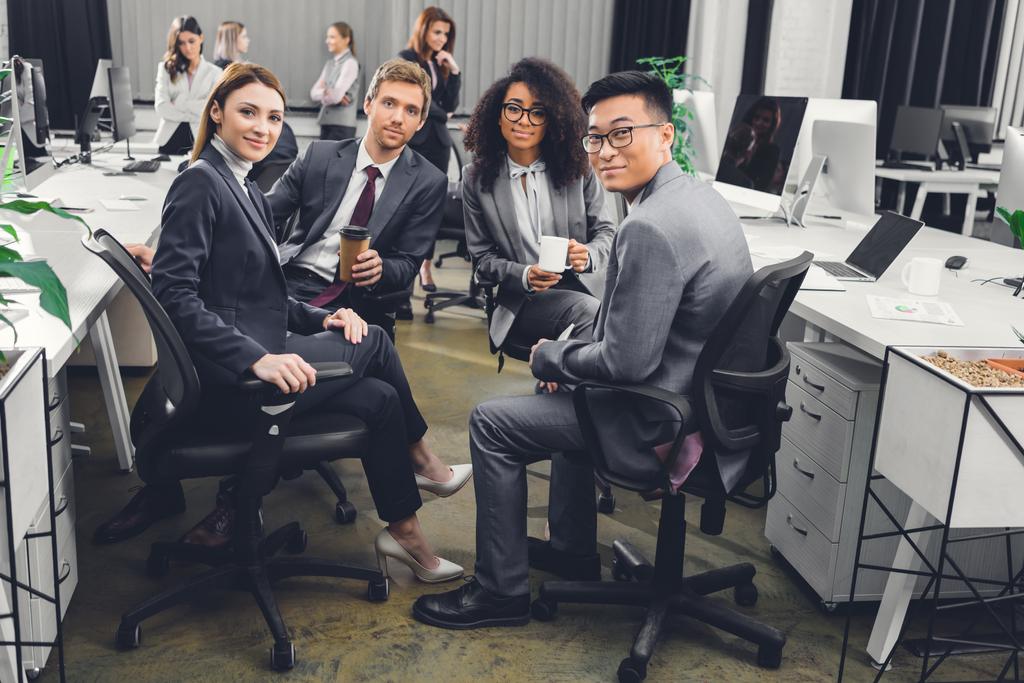 jovens empresários multiétnicos em vestuário formal sentados juntos e sorrindo para a câmera no escritório
    - Foto, Imagem