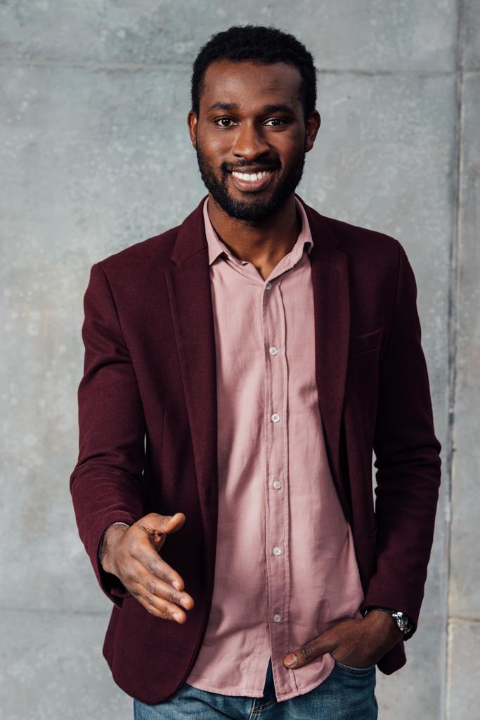 χαμογελώντας αφρικανικό αμερικανικό casual επιχειρηματίας outstretching το χέρι για χειραψία σε γκρίζο φόντο - Φωτογραφία, εικόνα