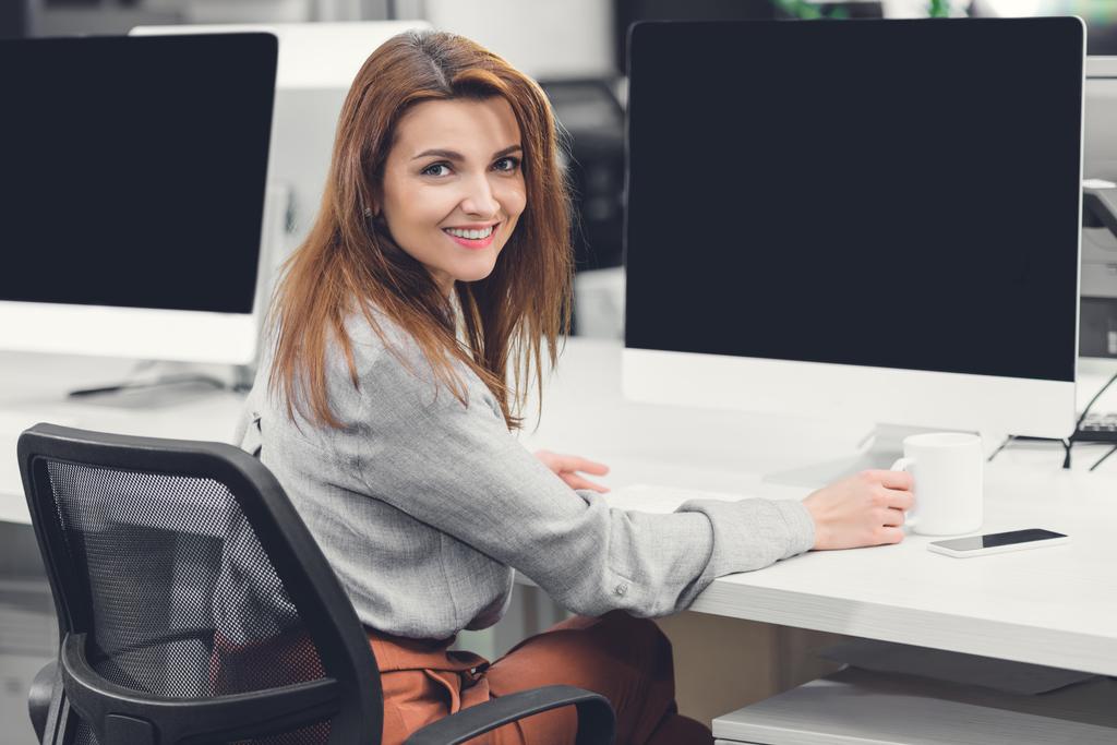 hermosa joven mujer de negocios que trabaja con la computadora de escritorio y sonriendo a la cámara en la oficina
 - Foto, Imagen