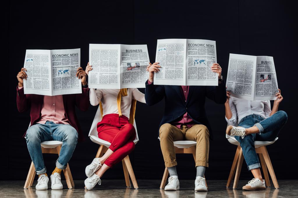 group of casual businesspeople with obscure faces sitting on chairs and reading business newspapers isolated on black - Photo, Image