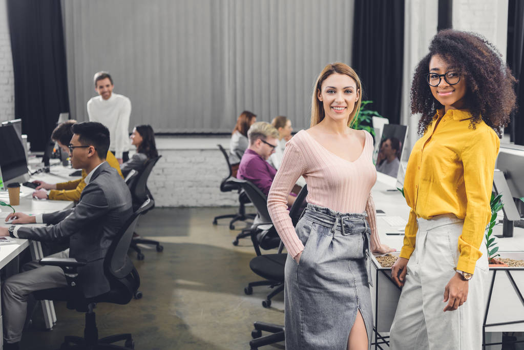 jóvenes empresarias multiétnicas sonriendo a la cámara mientras sus colegas trabajan detrás en la oficina
  - Foto, Imagen