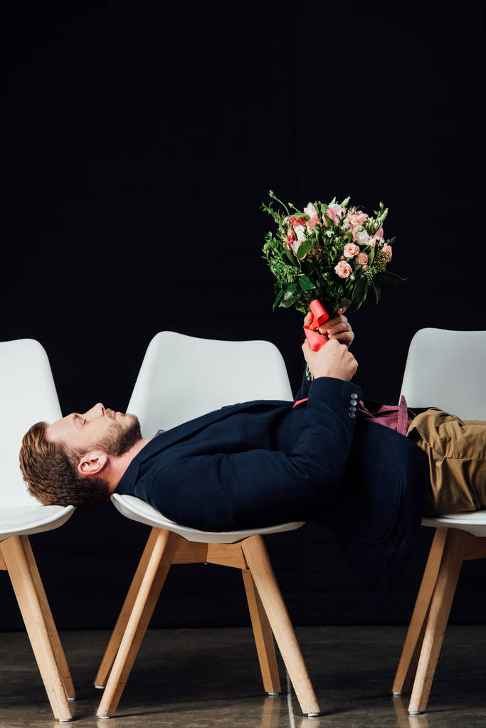 Mann mit geschlossenen Augen auf Stühlen liegend und Blumen isoliert auf schwarz haltend - Foto, Bild