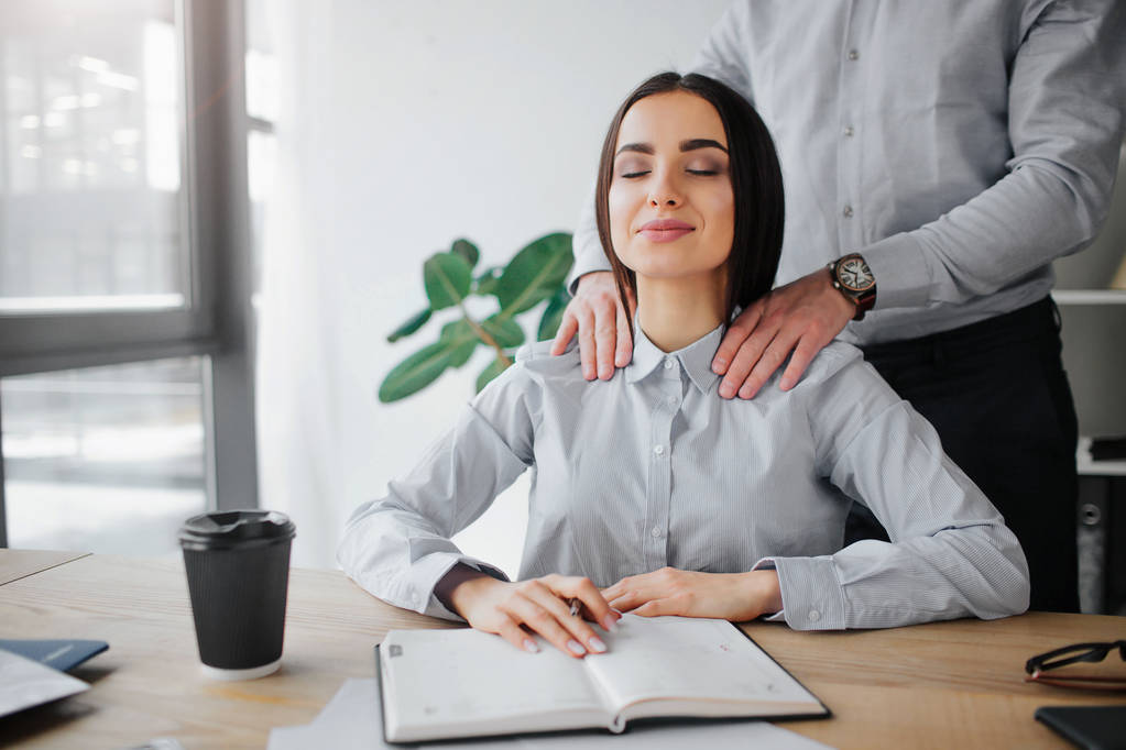 Young woman get attention from boss. He does shoulder massage for her. She is relaxed and pleased. Young woman sit in room at table. Guy stand behind her. - Photo, Image