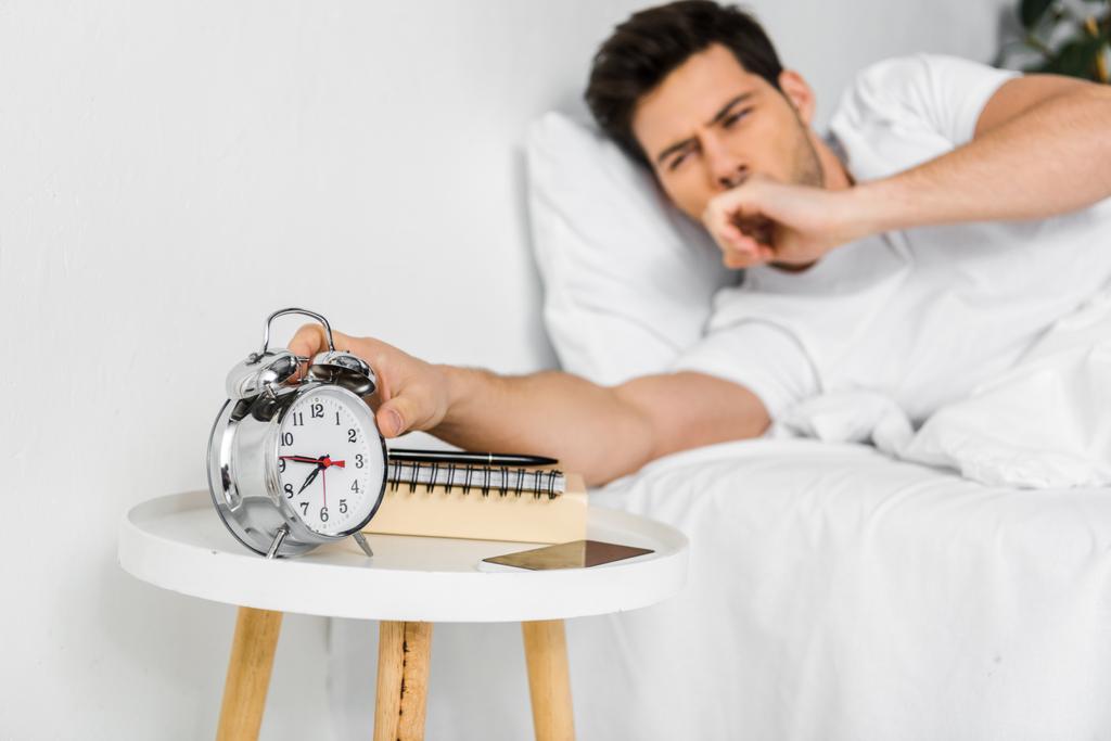 επιλεκτική εστίαση του ανθρώπου χασμουρητό ξυπνά και κοιτάζοντας το ρολόι συναγερμών  - Φωτογραφία, εικόνα